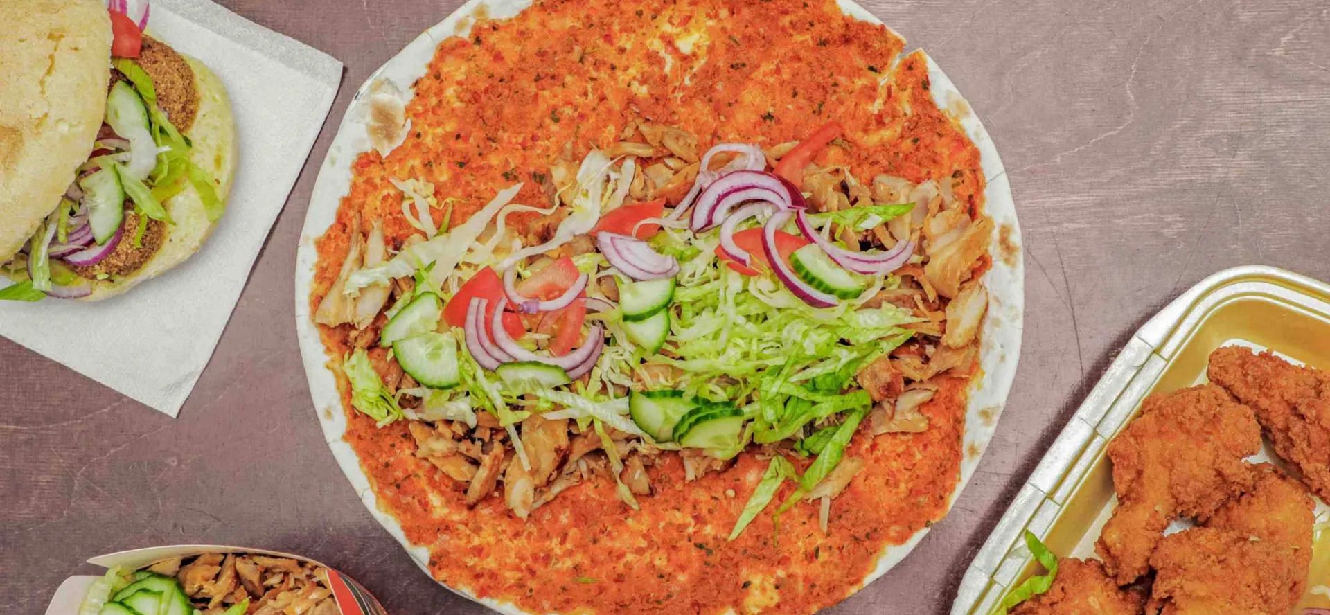 Bovenaanzicht van een houten tafel met een groot plat brood met vlees en salade, een broodje, een bak vlees met salade, twee sausjes en een bakje krokante kip.